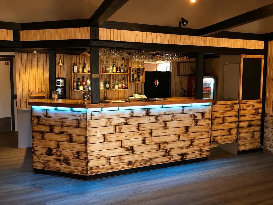 The bar at Fish House, faced in scorched timber planks lit from beneath, with glasses hanging above the counter and bottles on the shelves behind